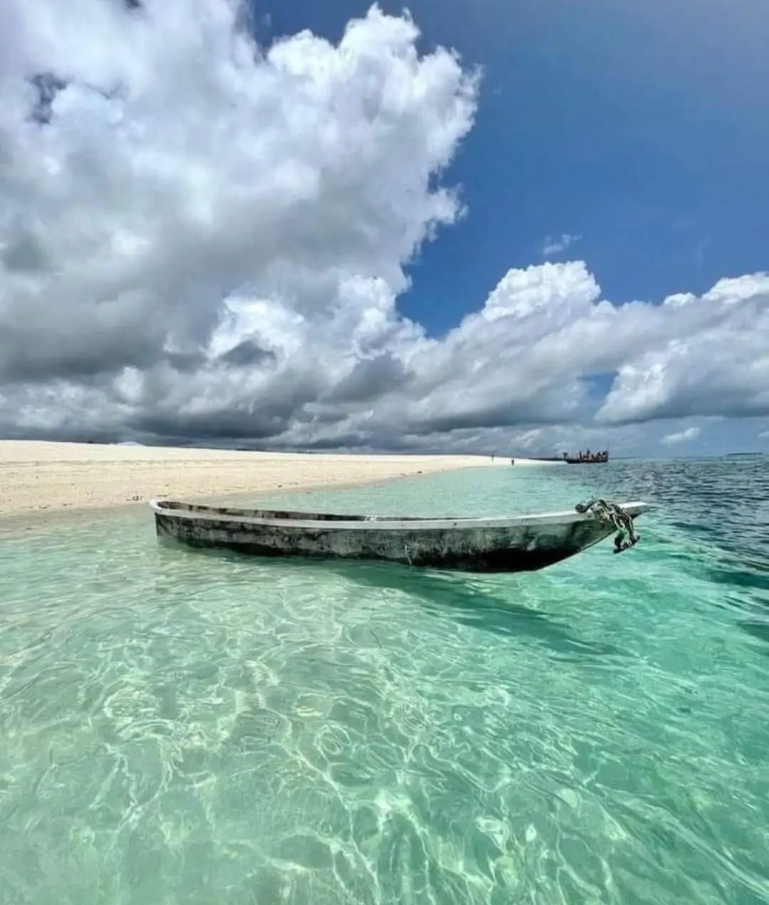 A wooden boat floating in glass-clear shallow water at the edge of a white sandbank, under tall cloud.