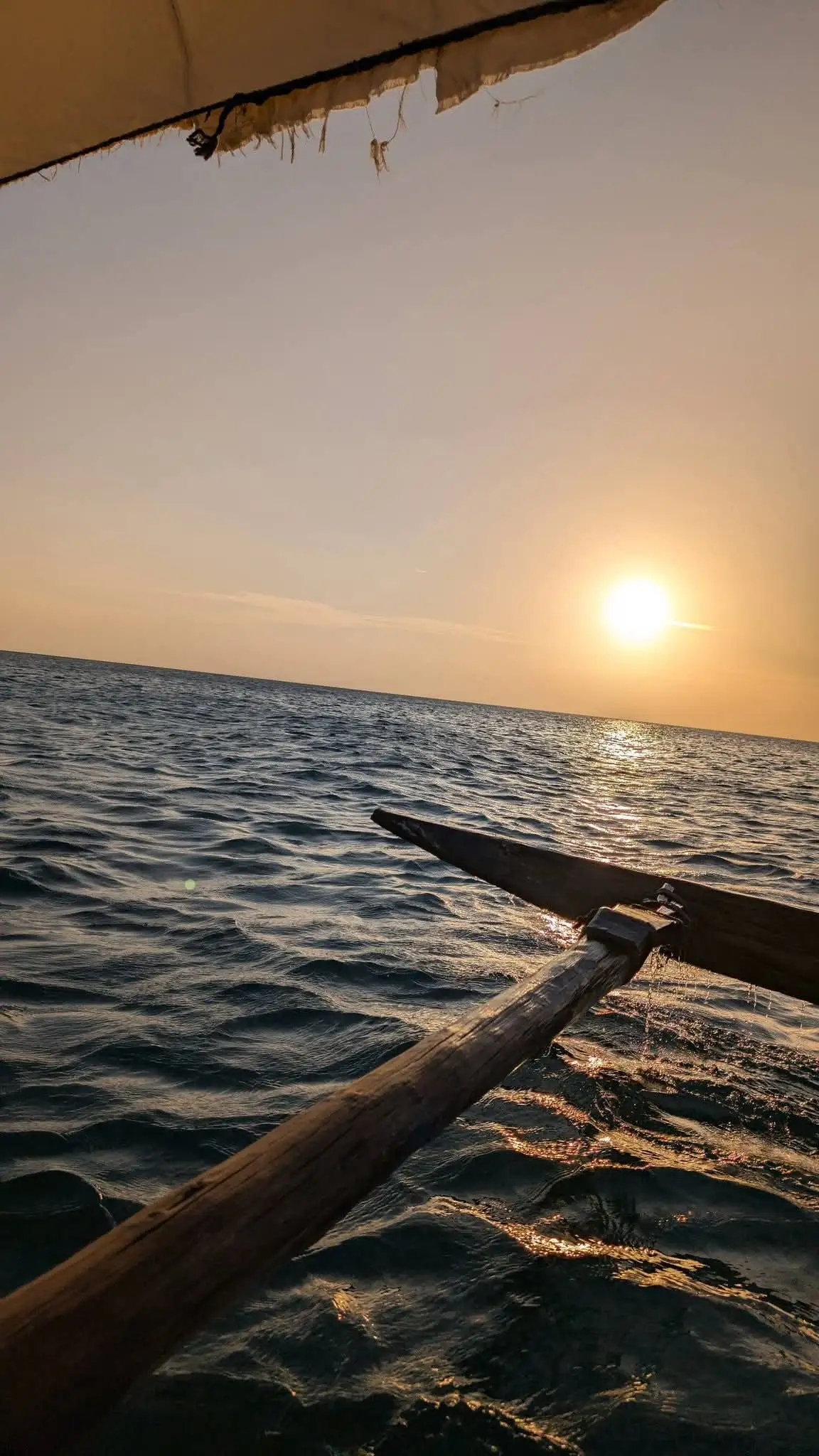The sun low over open water, seen past the wooden outrigger of a sailing dhow.