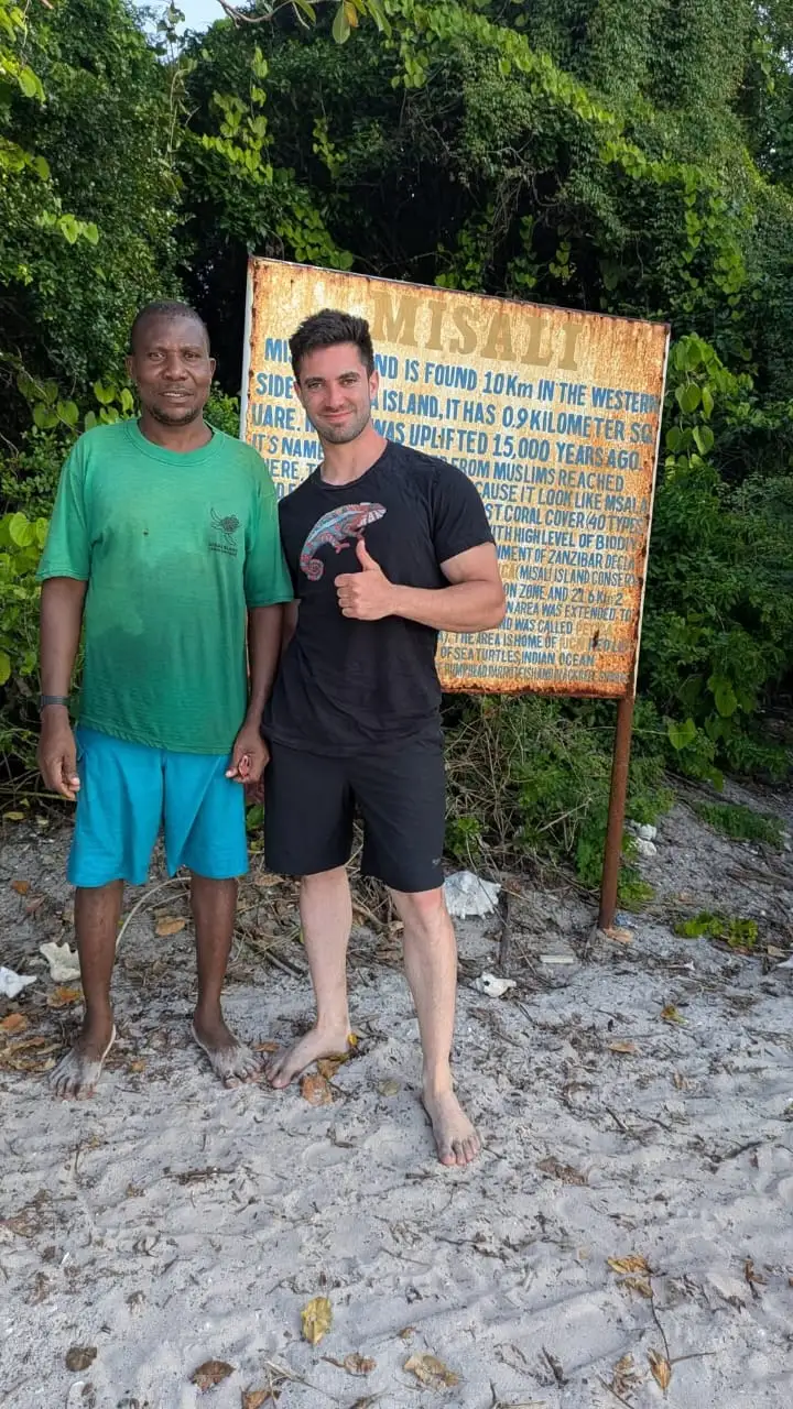 A guide and a guest standing barefoot on the sand beside the painted Misali Island information board.