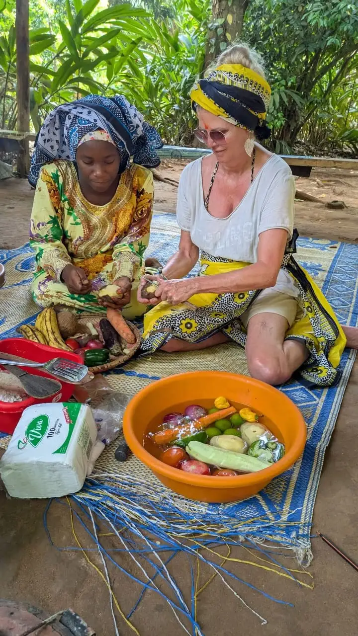 A guest and a local cook sitting on a woven mat, peeling vegetables into a bowl of tomatoes, limes and carrots.