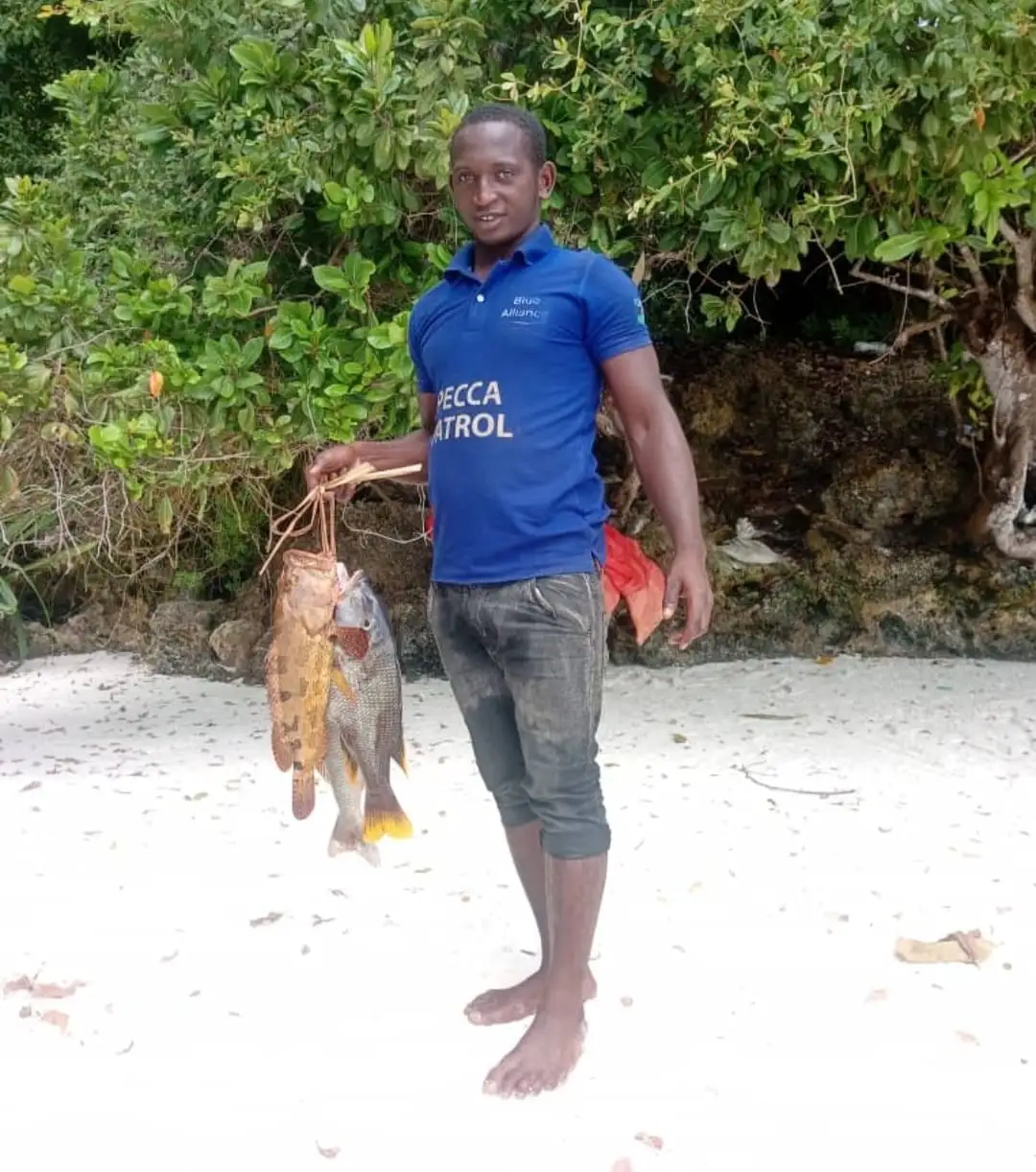 Two reef fish hanging from a line, held up on white sand in front of dense green bush.